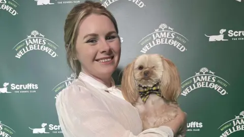 Lauraine Maddox A woman smiling in a white shirt, holding a one-eye shih-tzu dog wearing a Cornish bow tie, in front of a board saying 'Scruffts'