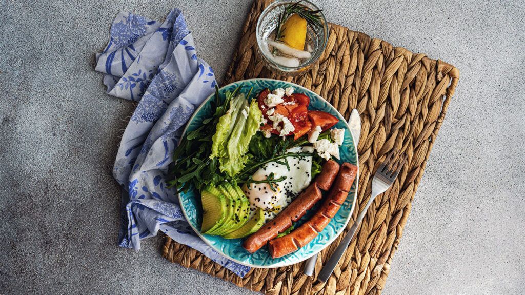 A plate of avocado, sausage, eggs, greens, and tomatoes, with lemon water on the side.