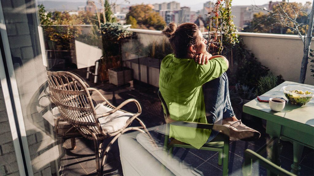A person sat on a balcony, in the sun, with a bowl of salad on the table next to them