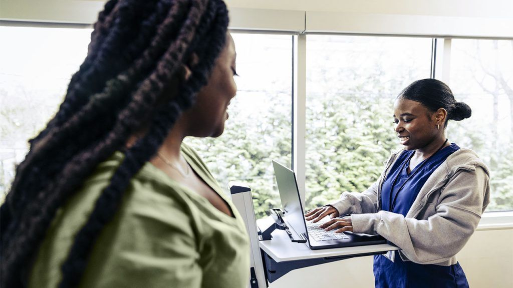 An oncology nurse takes notes on a laptop as a patient prepares for a mammogram to screen for breast cancer