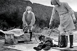 Photo en extérieur, deux hommes en costume médical entourent un homme au sol et l'aspergent d'un produit.