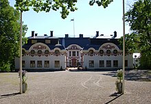 L'avant du château, avec une grande place de gravier et deux poteaux. Le château a des murs blancs et un toit bleu, et la façade est ornée de fenêtres carrées en bas et rondes à l'étage. Le portail en fer forgé donne sur une petite cour intérieure au fond de laquelle on voit une grande porte en bois.