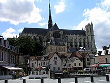 Seitliche Farbfotografie einer gotischen Kathedrale mit alten Gebäuden und Außenterrassen am vorderen Platz. Schmale Ziertürme sind an der ganzen Fassade und in der Mitte steht ein Anbau mit großem Kirchenfenster. In der Mitte des Kirchenschiffs befindet sich eine Dachlaterne mit vielen Verzierungen und rechts ein Eckturm.