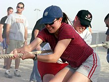 En la imagen se observa en el centro a una mujer de 20 a 30 años, vestida con ropa deportiva, con un short gris con dos rayas rojas en el centro y una remera roja, tomando con sus dos manos una soga grande y tirando la soga hacia el centro de su cuerpo, en el juego de la cinchada. Alrededor de ella, hay dos hombres como espectadores, y detrás de ella, hay otros dos hombres que la ayudan a tirar de la soga.