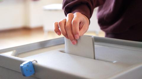 Eine Hand steckt einen Umschlag in eine versiegelte Wahlurne